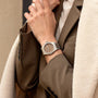 Person wearing a brown blazer and beige coat, displaying a Girard-Perregaux Laureato 38 with a silver case, brown dial, and octagonal bezel on their left wrist.