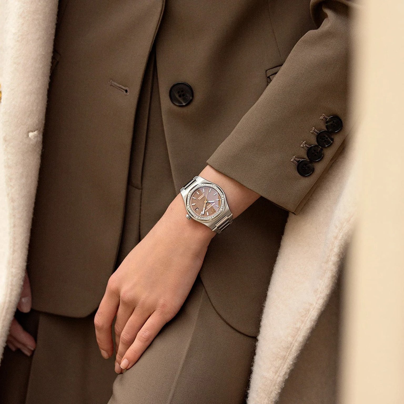 A person in a brown suit and cream coat is shown from the waist down, wearing a Girard-Perregaux Laureato 38 with an octagonal bezel on their left wrist.