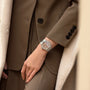A person in a brown suit and cream coat is shown from the waist down, wearing a Girard-Perregaux Laureato 38 with an octagonal bezel on their left wrist.