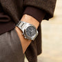 A person dressed in brown and gray wears a Girard-Perregaux Laureato Chronograph Ti49 in Grade 5 titanium with a black dial, their hand partially in a pocket.