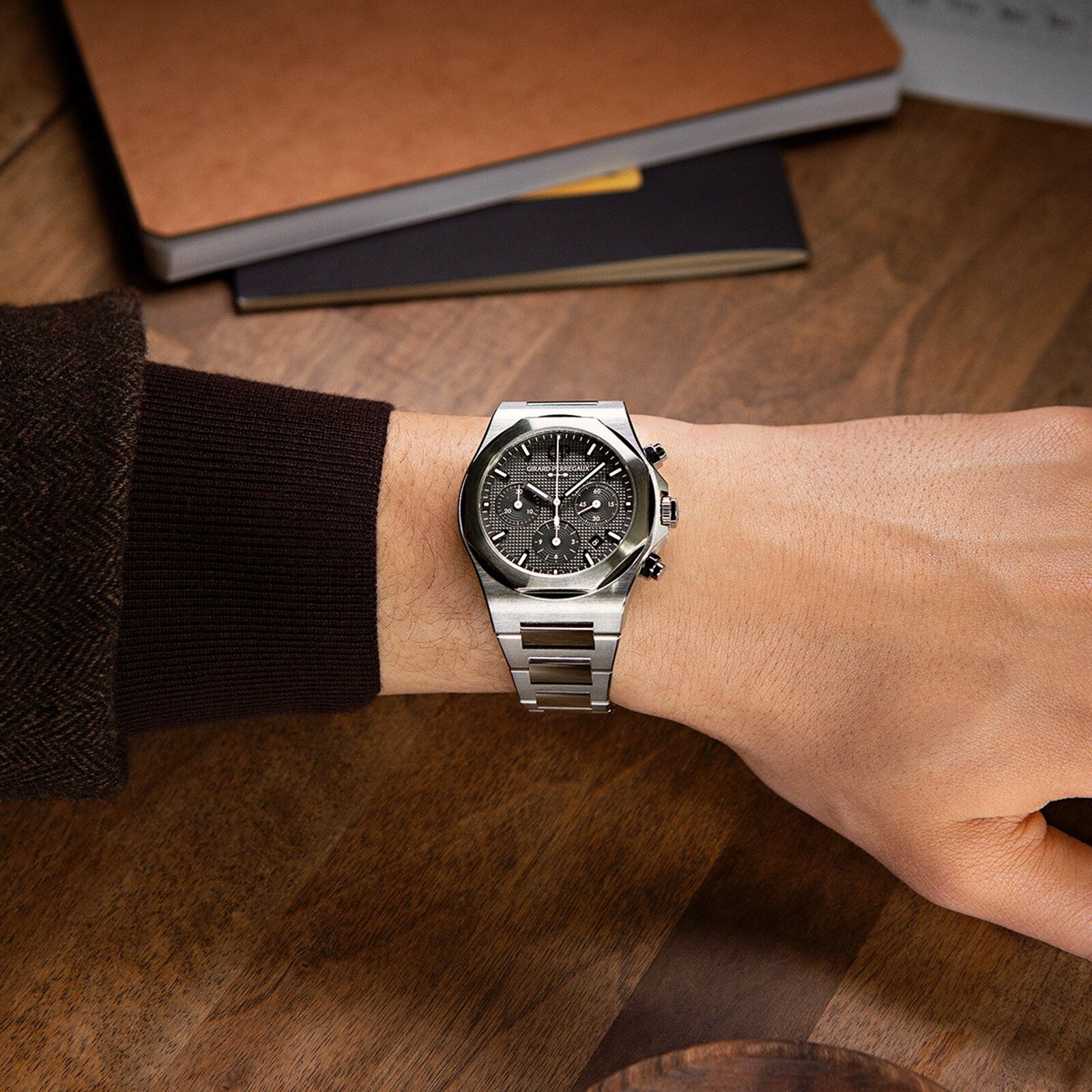 A person wearing a Girard-Perregaux Laureato Chronograph Ti49 with a black chronograph dial rests their arm on a wooden table beside notebooks.