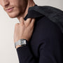 A man in a black sweater holds a jacket over his shoulder, wearing a Cartier Santos de Cartier silver wristwatch with a square steel case and Roman numeral markers.
