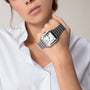 Wearing a white shirt, a person showcases the Cartier Santos de Cartier wristwatch, featuring a square steel case and Roman numerals, while holding their wrist near their chin.