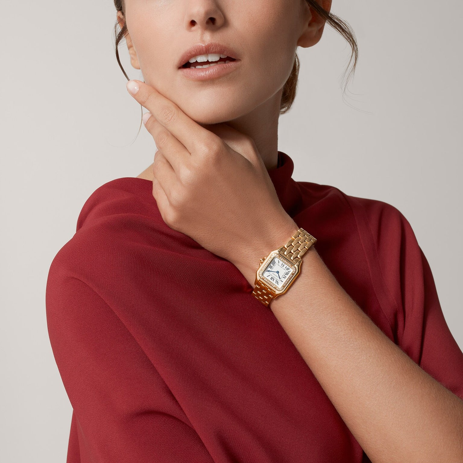 A woman in a red top wears the Panthère de Cartier watch by Cartier, posing with her hand touching her face against a neutral background.