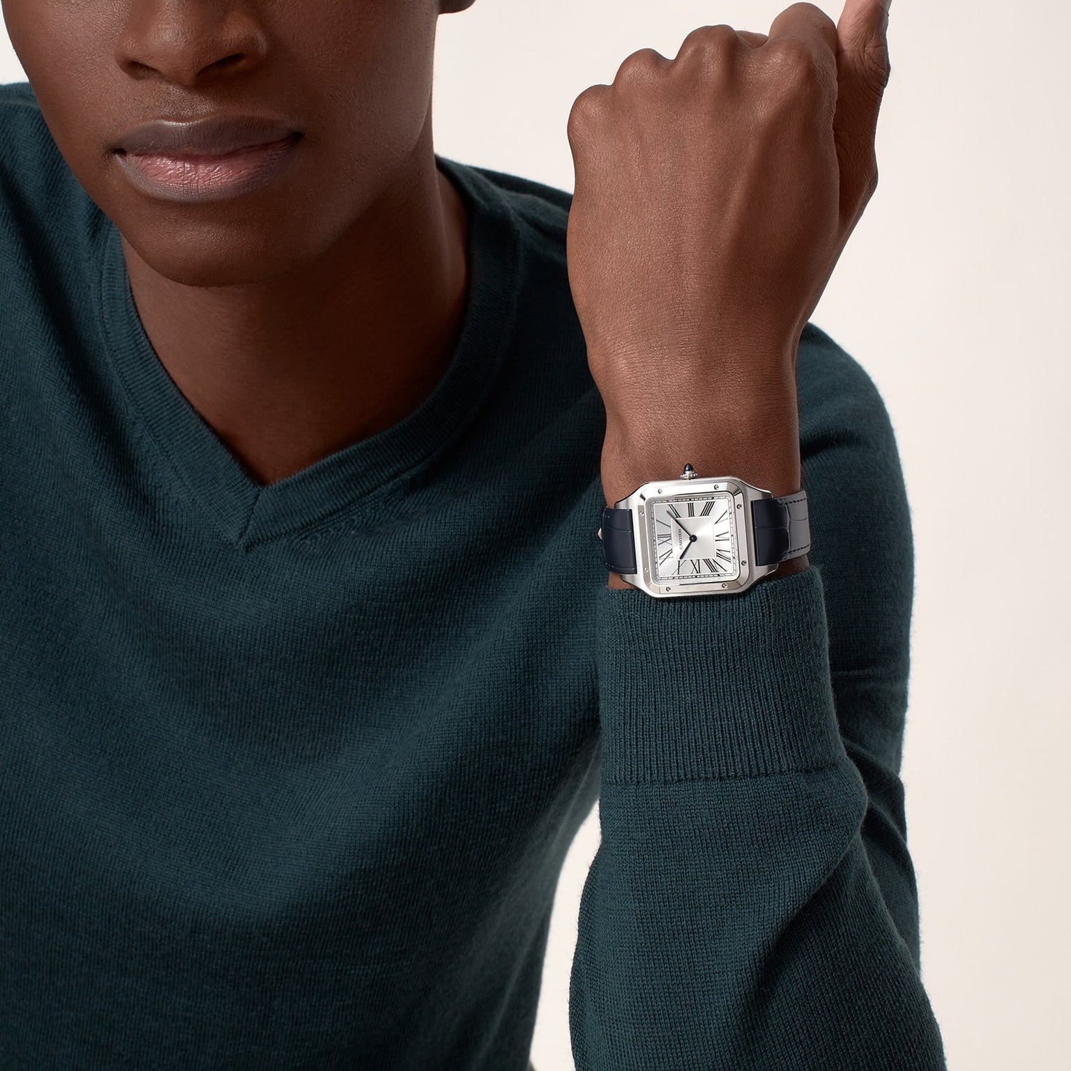 A person in a dark green sweater showcases the Cartier Santos-Dumont watch, which has a square face, mechanical movement, Roman numeral markers, and a black leather strap.