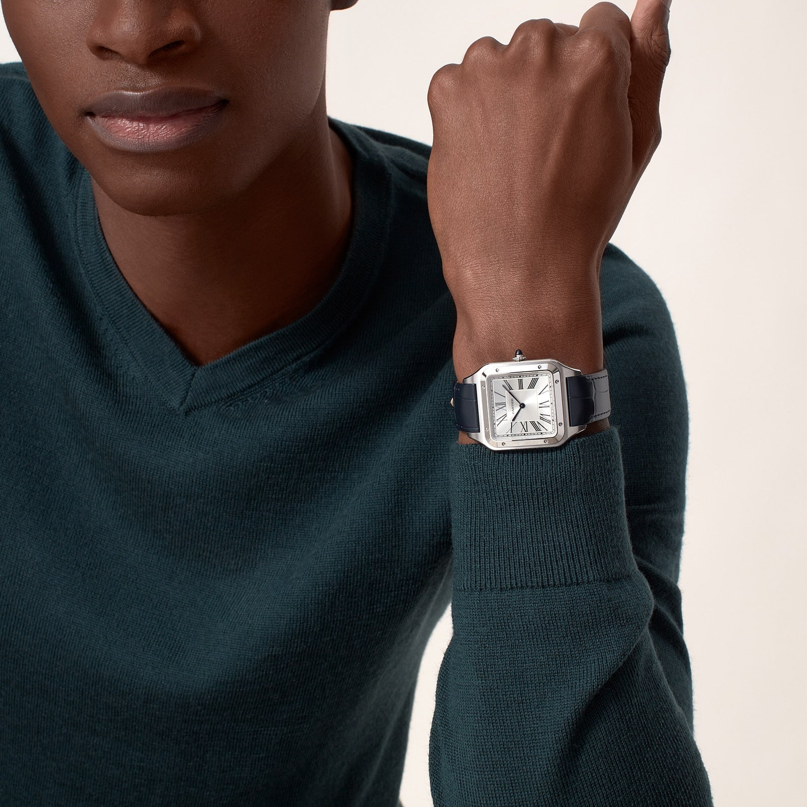 A person in a dark green sweater showcases the Cartier Santos-Dumont watch, which has a square face, mechanical movement, Roman numeral markers, and a black leather strap.