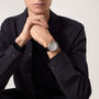 A person in a dark jacket wears a sophisticated Ballon Bleu de Cartier watch by Cartier, sitting with hands clasped under their chin and facing forward against a plain background.