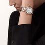 A person wearing the Ballon Bleu de Cartier watch, featuring a two-tone metal bracelet with rose gold, a round white dial with black Roman numerals, and a crown guard, paired with a black long-sleeve top.