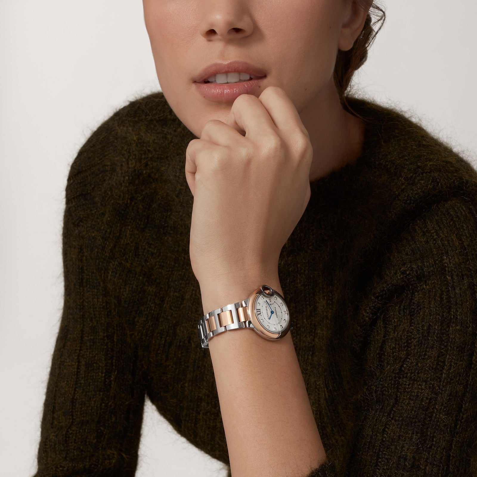 A person in a dark green sweater rests their chin on their hand, showcasing a Cartier Ballon Bleu de Cartier watch with a round silver face and guilloché dial.