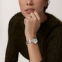 A person in a dark green sweater rests their chin on their hand, showcasing a Cartier Ballon Bleu de Cartier watch with a round silver face and guilloché dial.