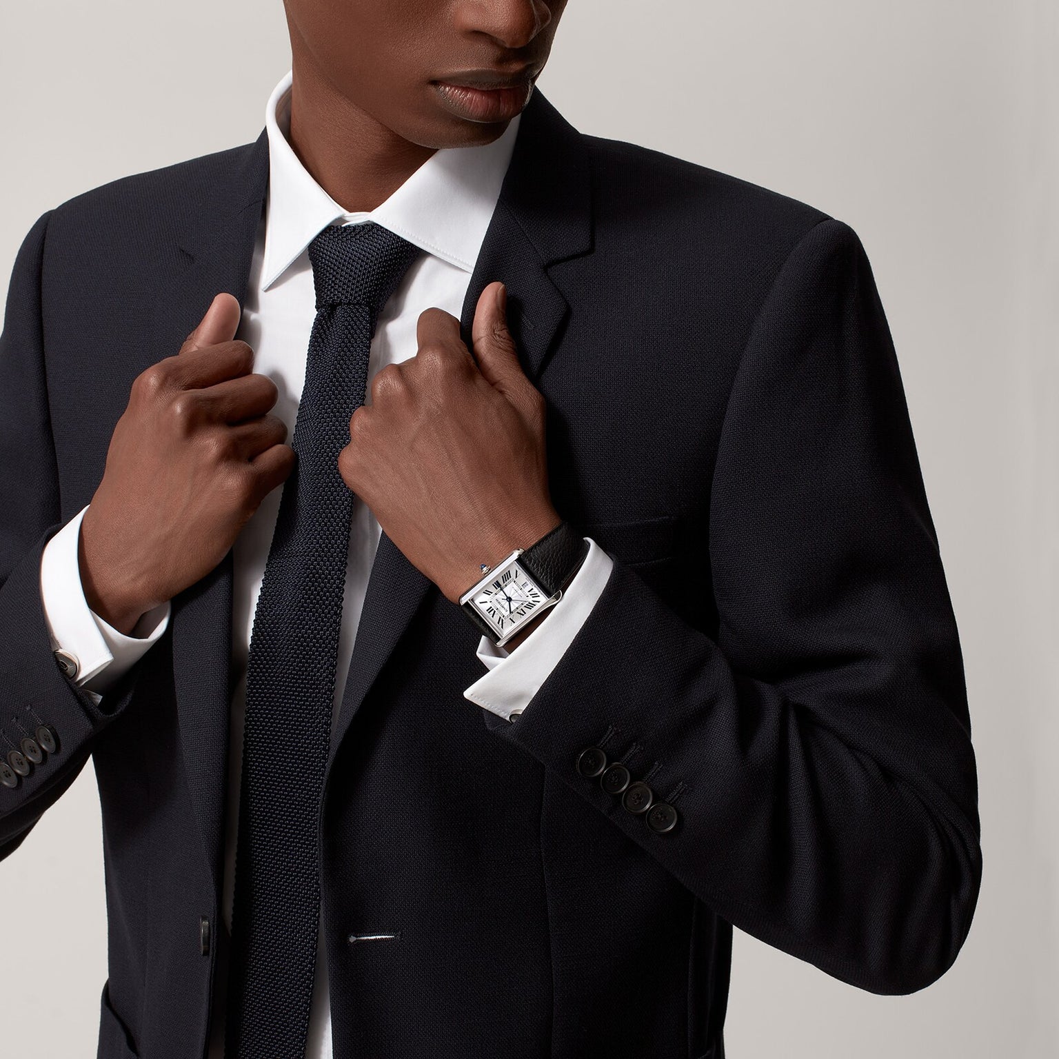 A man in a dark suit and tie adjusts his jacket, revealing the Cartier Tank Must de Cartier watch with a silvered flinqué dial and black calfskin strap on his left wrist against a plain background.