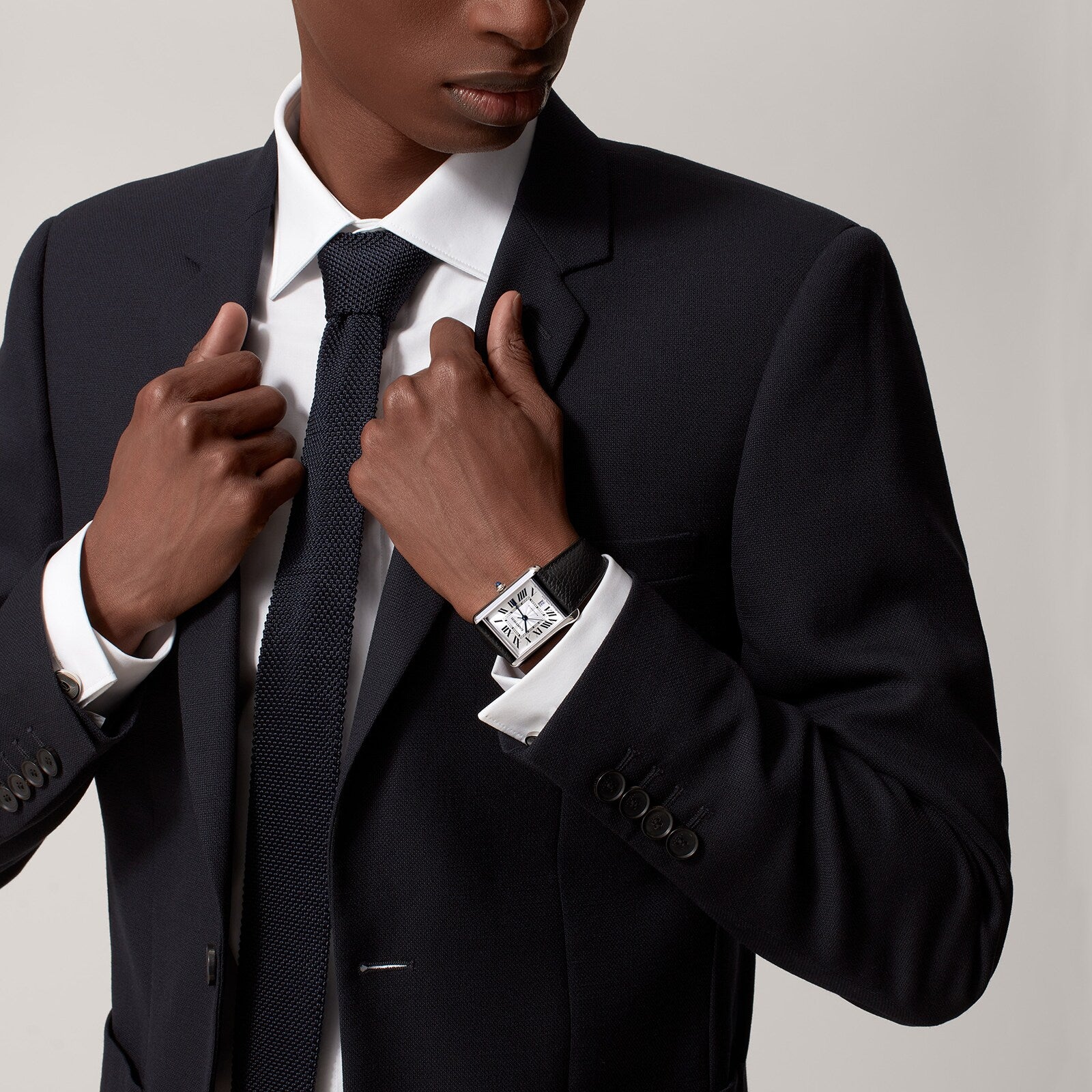 A man in a dark suit and tie adjusts his jacket, revealing the Cartier Tank Must de Cartier watch with a silvered flinqué dial and black calfskin strap on his left wrist against a plain background.