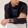 A man in a dark jacket and denim shirt rests his arms on a white surface, wearing the Cartier Ballon Bleu de Cartier with a striking blue guilloché dial on his left wrist.