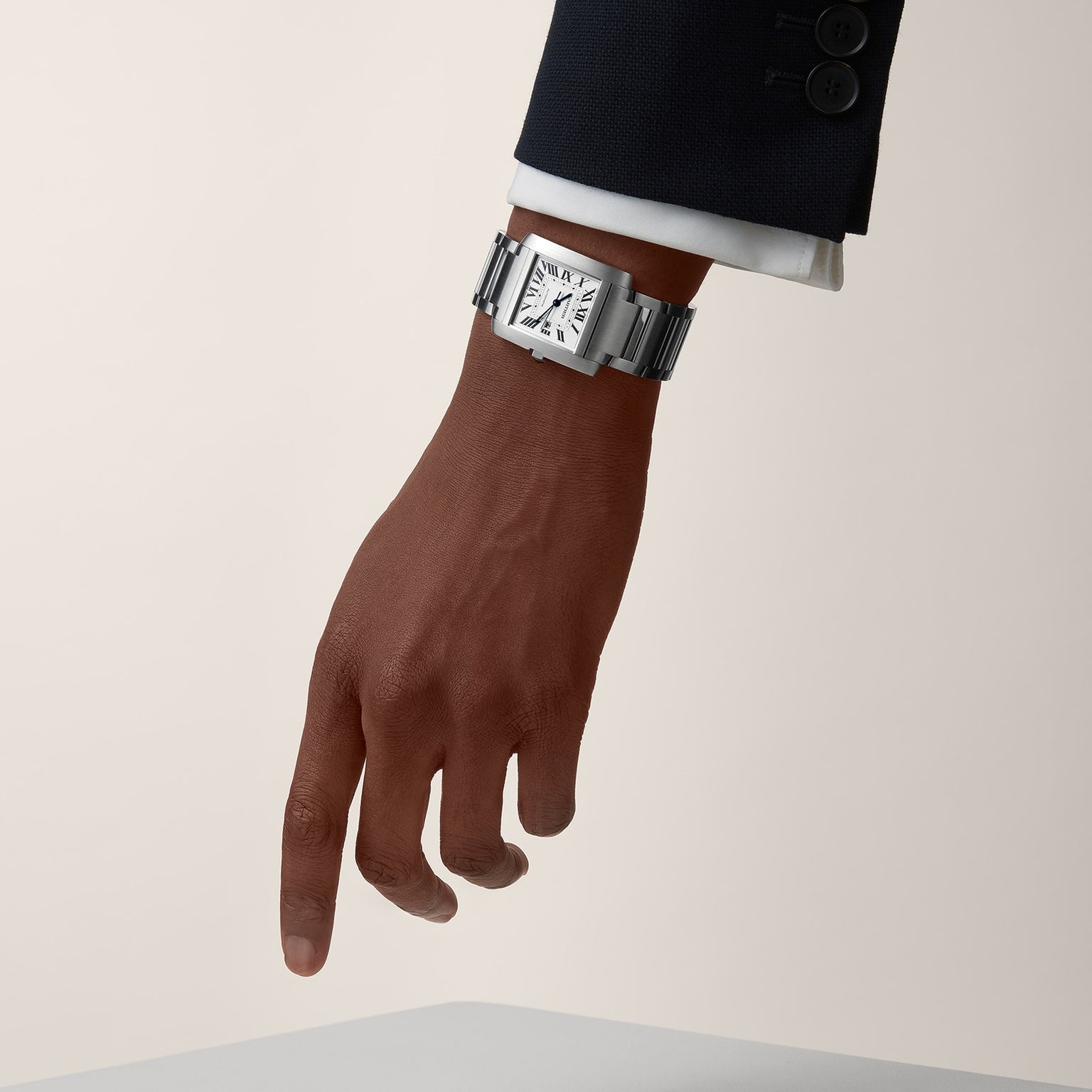 A person in a dark suit points downward, wearing a Cartier Tank Française watch with a steel bracelet and a white dial featuring Roman numerals.