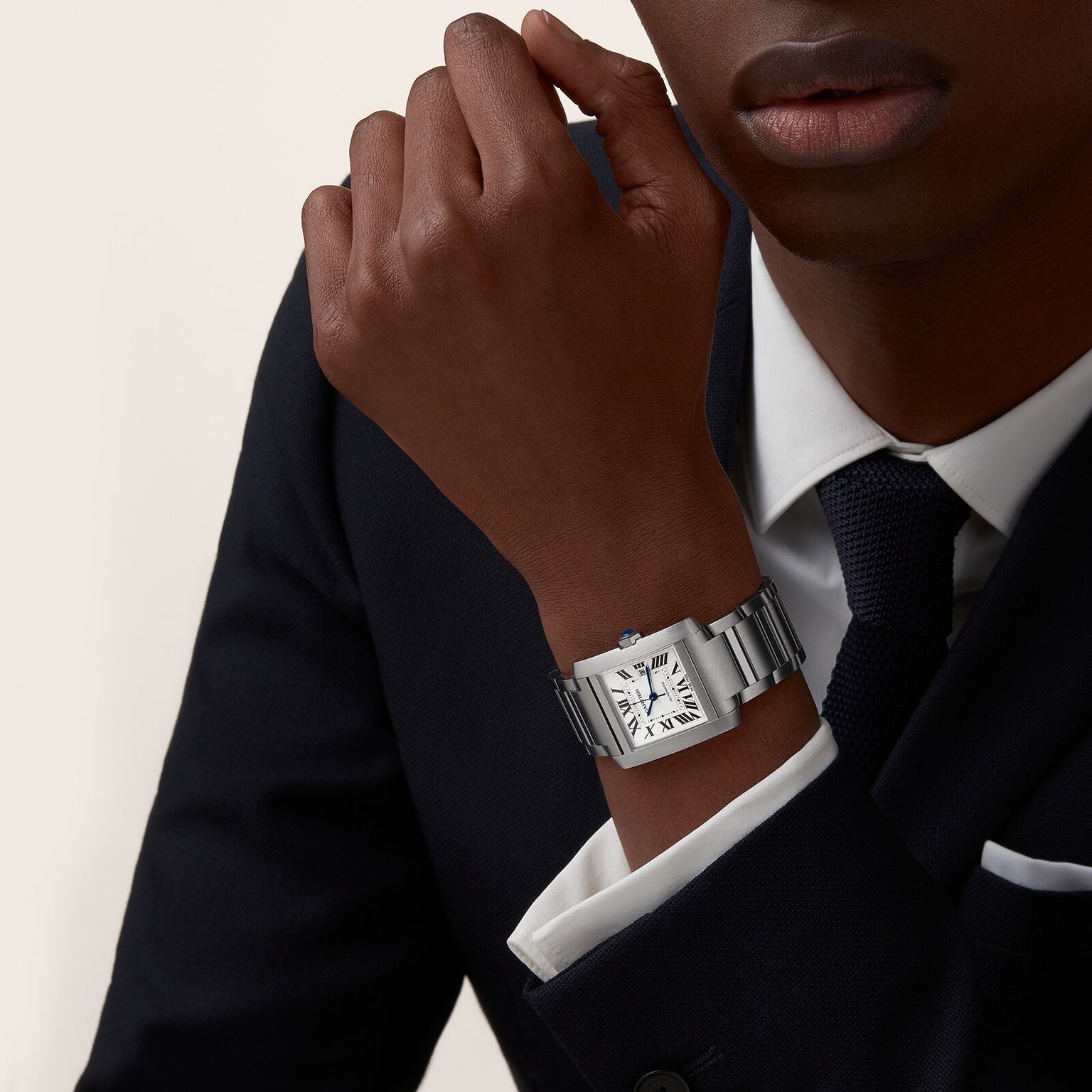 A person in a dark suit and tie rests their chin on their hand, wearing a Cartier Tank Française watch with a white dial, Roman numerals, and a polished steel bracelet.