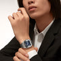 A person in a black blazer and white shirt showcases a Cartier Santos de Cartier wristwatch with a blue square face and square steel case on their left wrist.