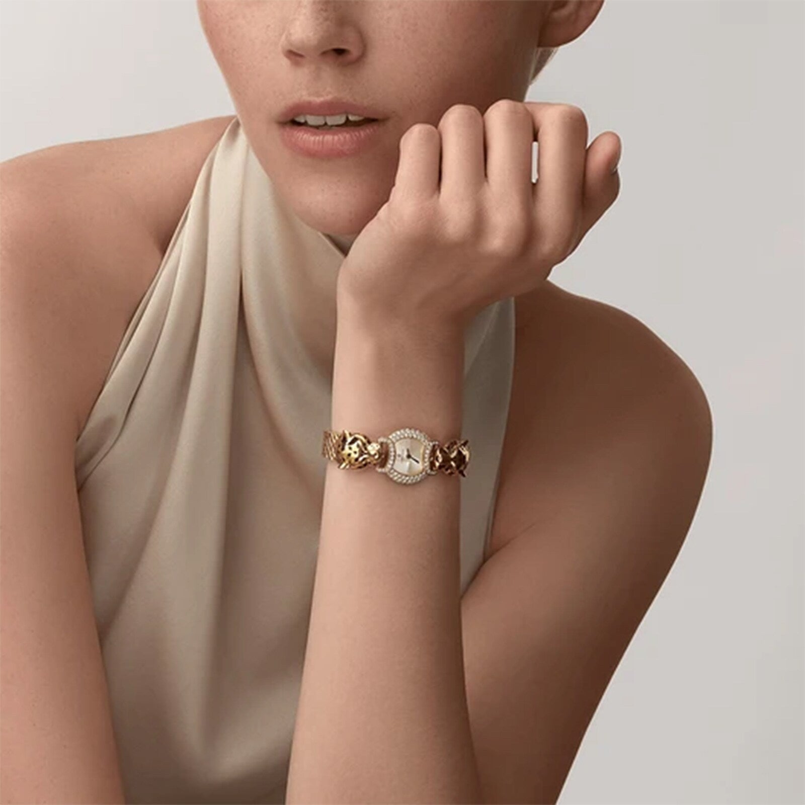 A woman in a sleeveless, light top rests her chin on her hand, showcasing Cartier’s La Panthère de Cartier yellow gold watch featuring panther heads and brilliant-cut diamonds on its decorative band.