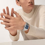 A person in a cream turtleneck showcases the Cartier Santos de Cartier aviation watch with a blue dial, resting their arms on a light surface.