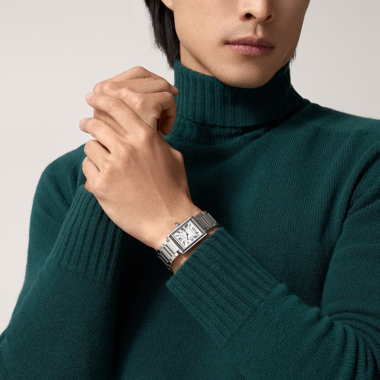 A person wearing a green turtleneck and the Cartier Tank Must de Cartier wristwatch—a silver rectangular timepiece with a white face, steel case, and quartz movement—clasps their hands together.