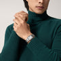 A person wearing a green turtleneck and the Cartier Tank Must de Cartier wristwatch—a silver rectangular timepiece with a white face, steel case, and quartz movement—clasps their hands together.