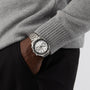 A person in a grey sweater and black pants has their hand in their pocket, displaying an OMEGA Speedmaster Racing wristwatch with a white chronograph dial.