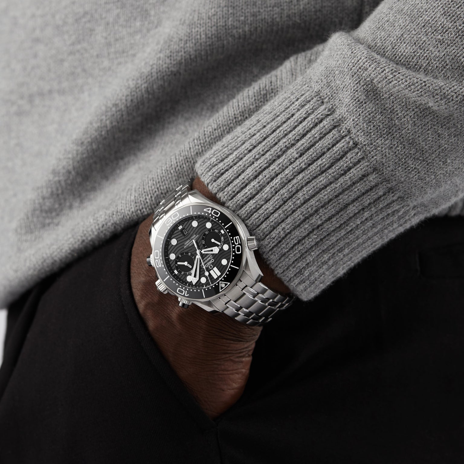 A person in a grey sweater and black pants has their hand in their pocket, showcasing an OMEGA Seamaster Diver 300M watch with a sleek black dial.