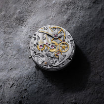 A close-up of OMEGA’s Speedmaster Moonwatch Professional reveals the intricate 3861 calibre movement with visible gears and screws, set against a textured gray surface.