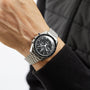 A person wears an OMEGA Speedmaster Moonwatch Professional with a black dial and three subdials, paired with a black quilted jacket.