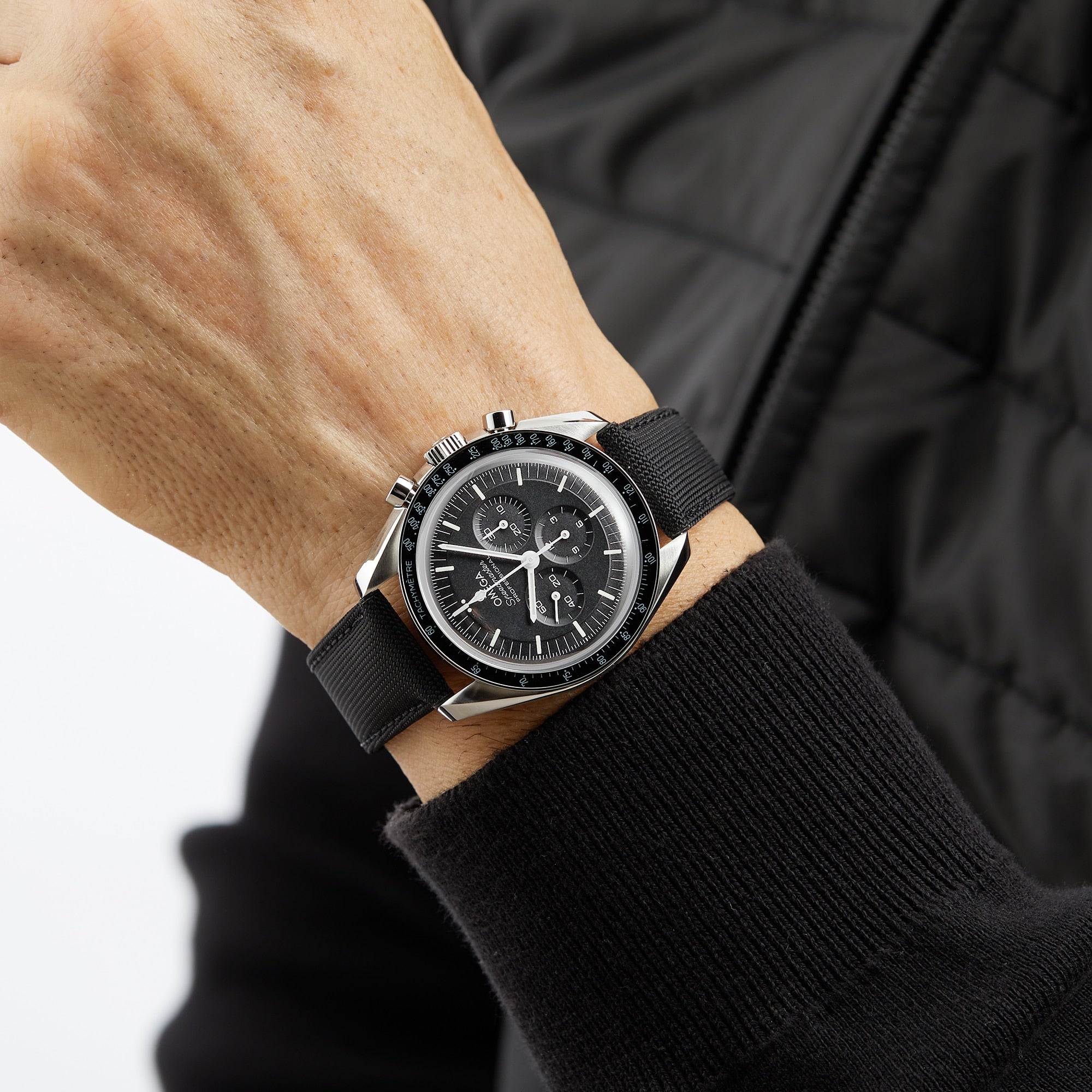 A person wearing an OMEGA Speedmaster Moonwatch Professional with a black strap and a black quilted jacket.