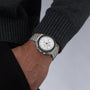 A person wearing an OMEGA Speedmaster Moonwatch Professional with a white dial and metal band, dressed in a dark sweater and posing with one hand in their pocket.