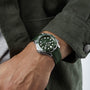 A person wears a green OMEGA Seamaster Diver 300M with a matching strap and jacket, their left hand on their right arm, highlighting the watchs precision and water resistance.
