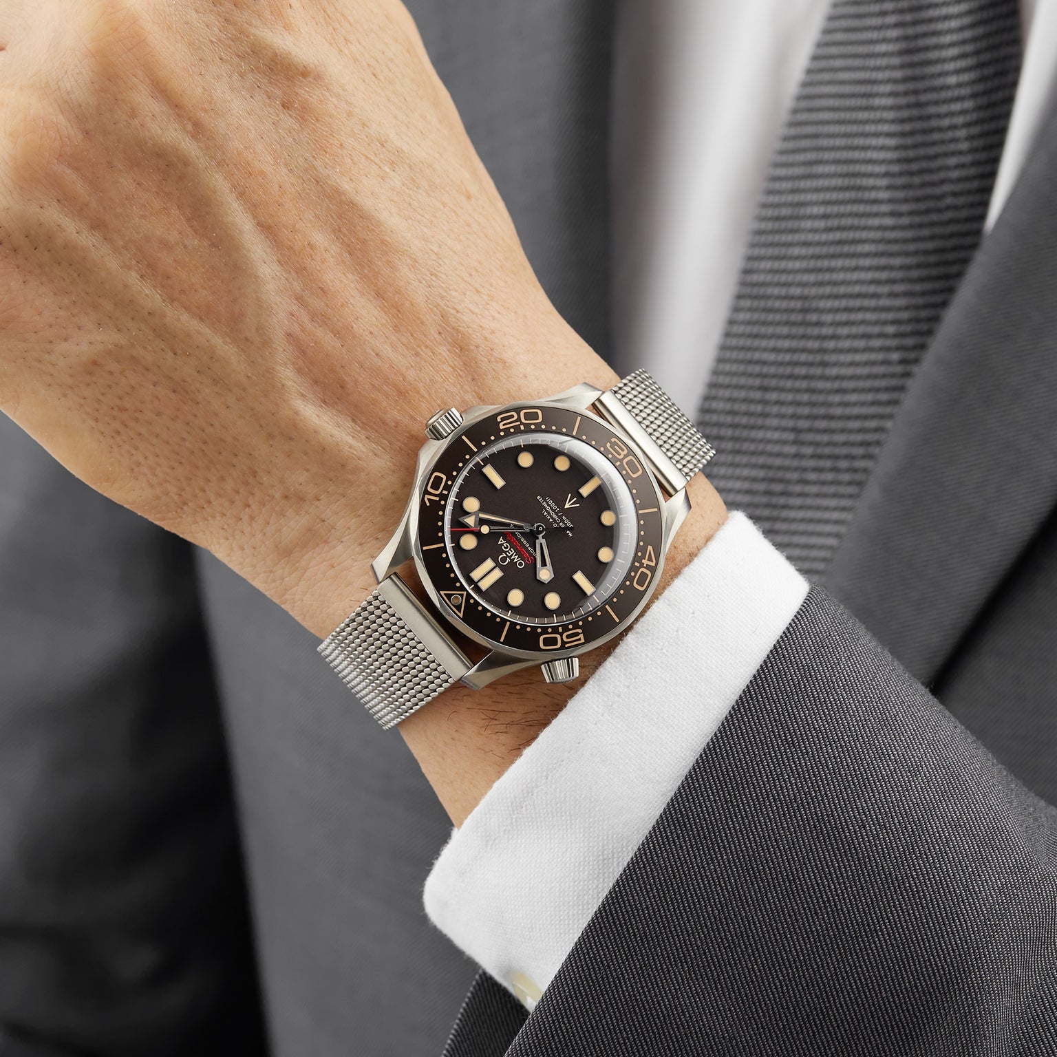 A person in a grey suit wears the OMEGA Seamaster Diver 300M James Bond 007 Edition with a black and gold bezel and mesh metal strap on their left wrist.