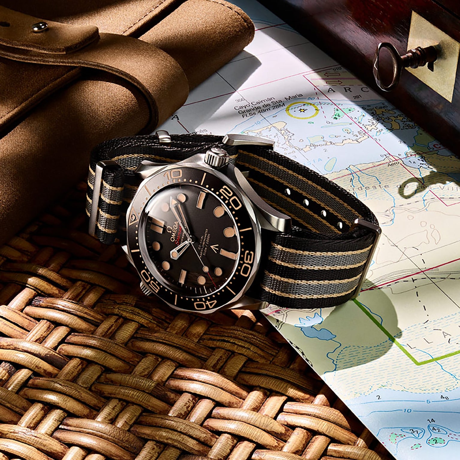 The OMEGA Seamaster Diver 300M James Bond 007 Edition with a black and tan NATO strap rests on a woven surface, beside a map, leather pouch, and wooden box.