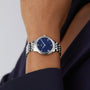An individual wearing an OMEGA De Ville Prestige with a stainless steel case, blue dial, and metal link band, paired with a dark long-sleeve outfit.