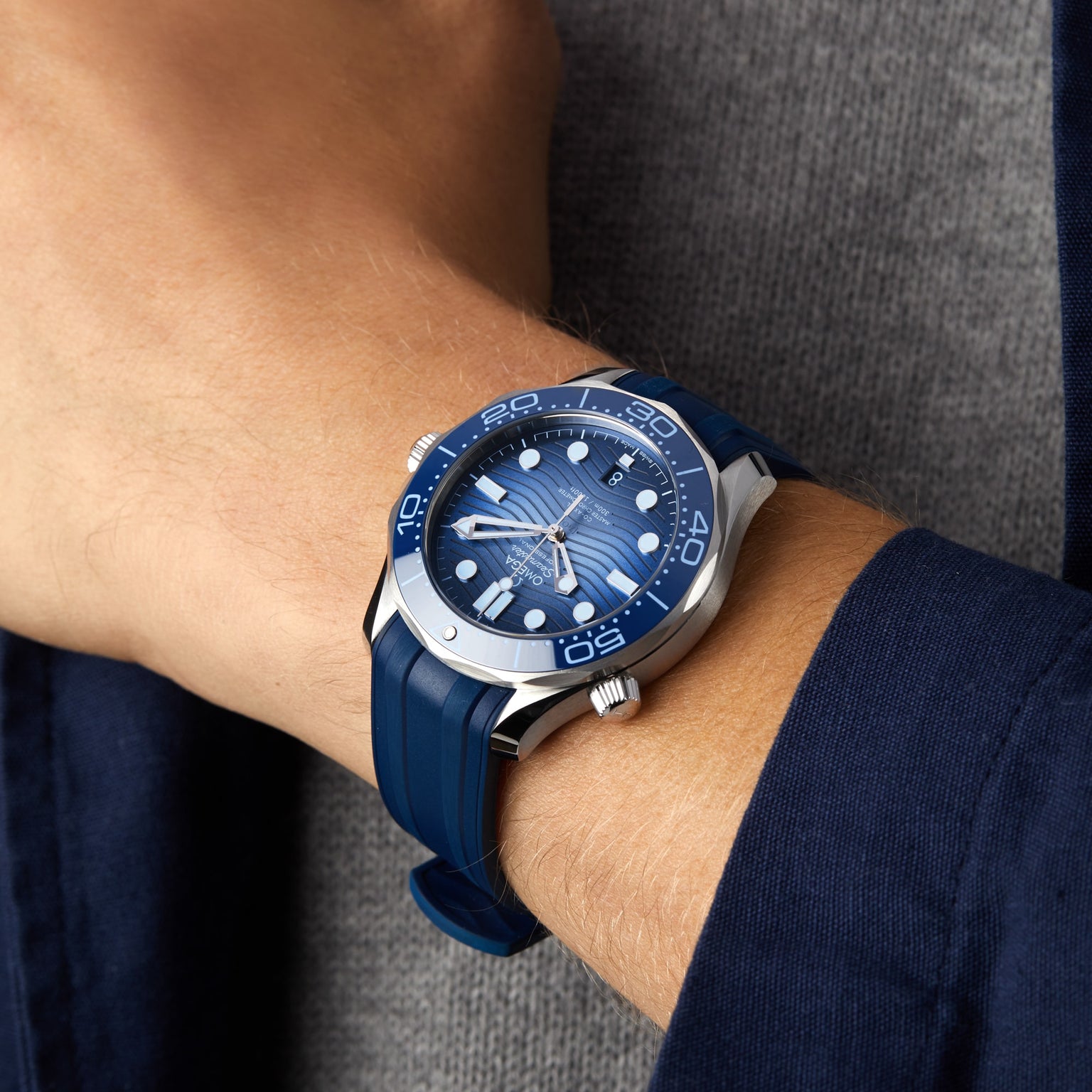 An individual sporting an OMEGA Seamaster Diver 300M Summer Blue with a silver bezel on their left wrist, paired with a grey shirt and navy blue jacket.