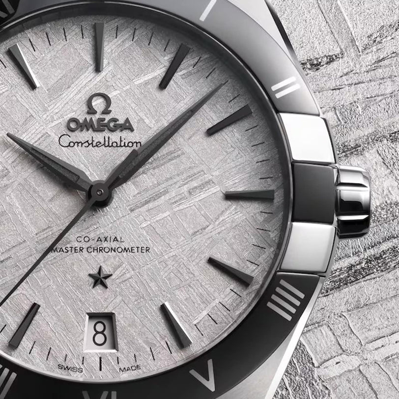 Close-up of an OMEGA Constellation watch featuring a textured silver dial, black hour markers, and a date window at 6 o’clock, set against a matching background.