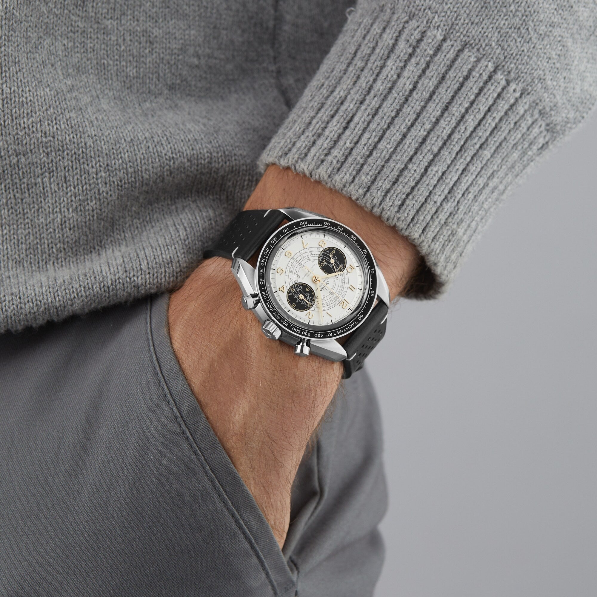 A person in a grey sweater and pants has their left hand in their pocket, showcasing an OMEGA Speedmaster Chronoscope Paris 2024 Olympics wristwatch with a black strap.