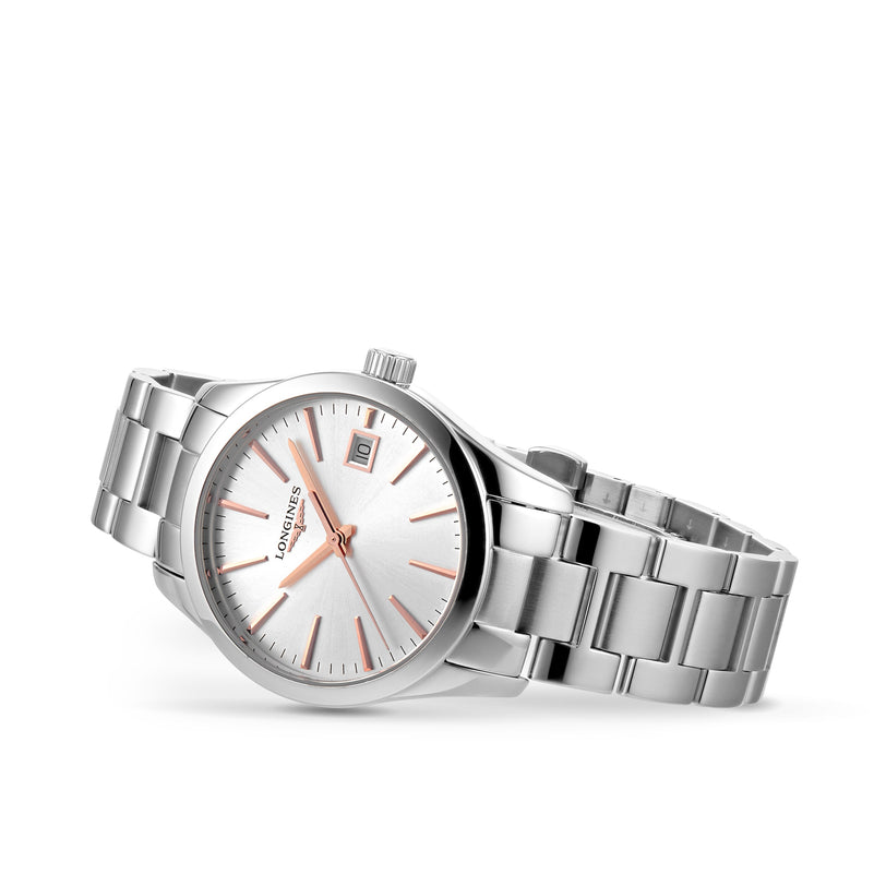 A Longines Conquest Classic silver wristwatch with a stainless steel case, metal band, silver dial, rose gold markers and hands, and a date window at 3 o’clock is shown on a white background.