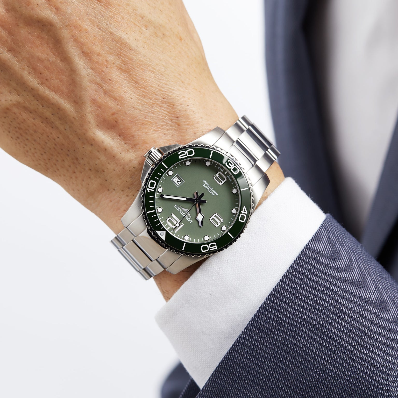 A person in a navy suit and white shirt wears a Longines HydroConquest diving watch with a silver metal band, green face, and white indices.