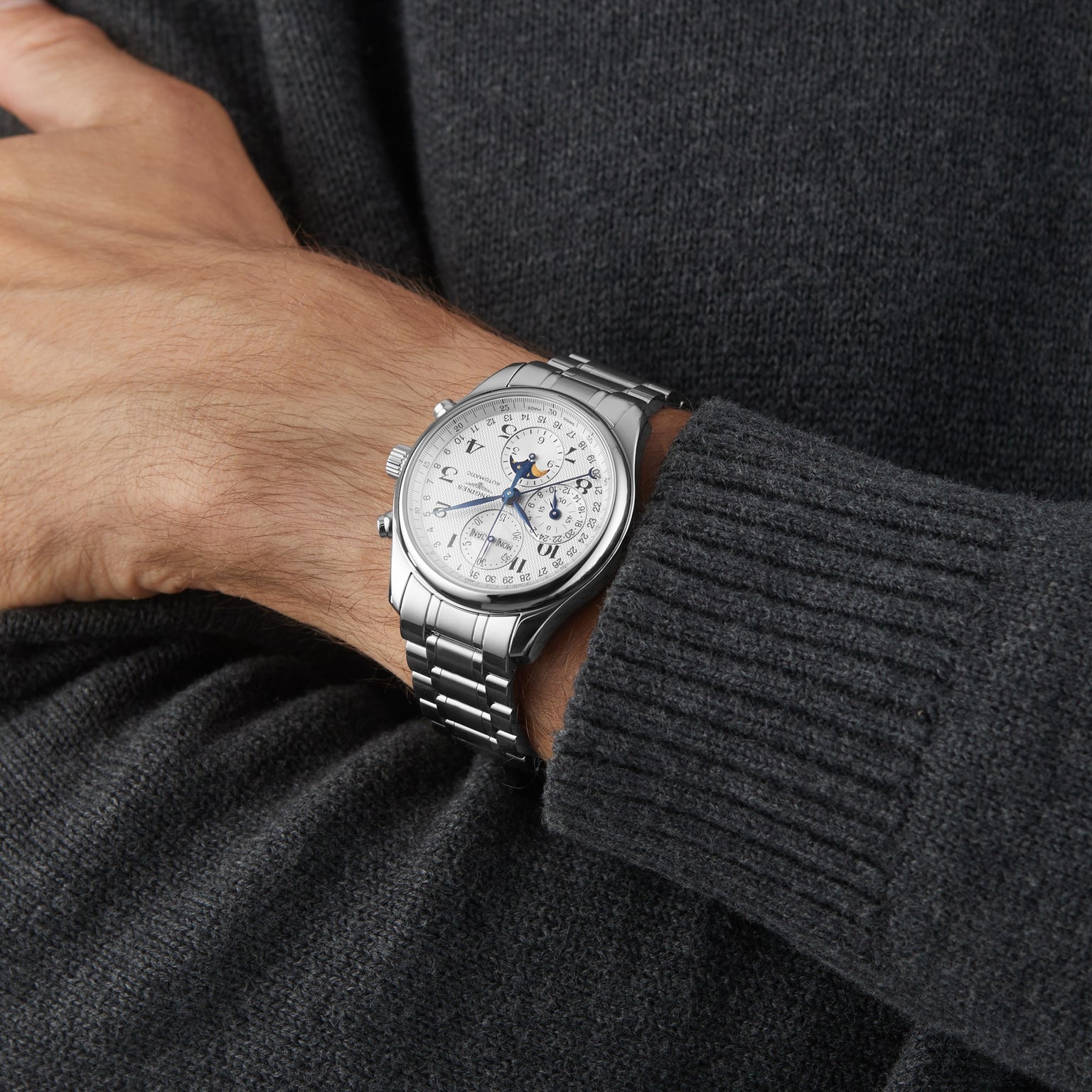 A person in a dark knit sweater wears a Longines Master Collection Chrono Moonphase silver analog wristwatch with a moonphase display and metal bracelet.