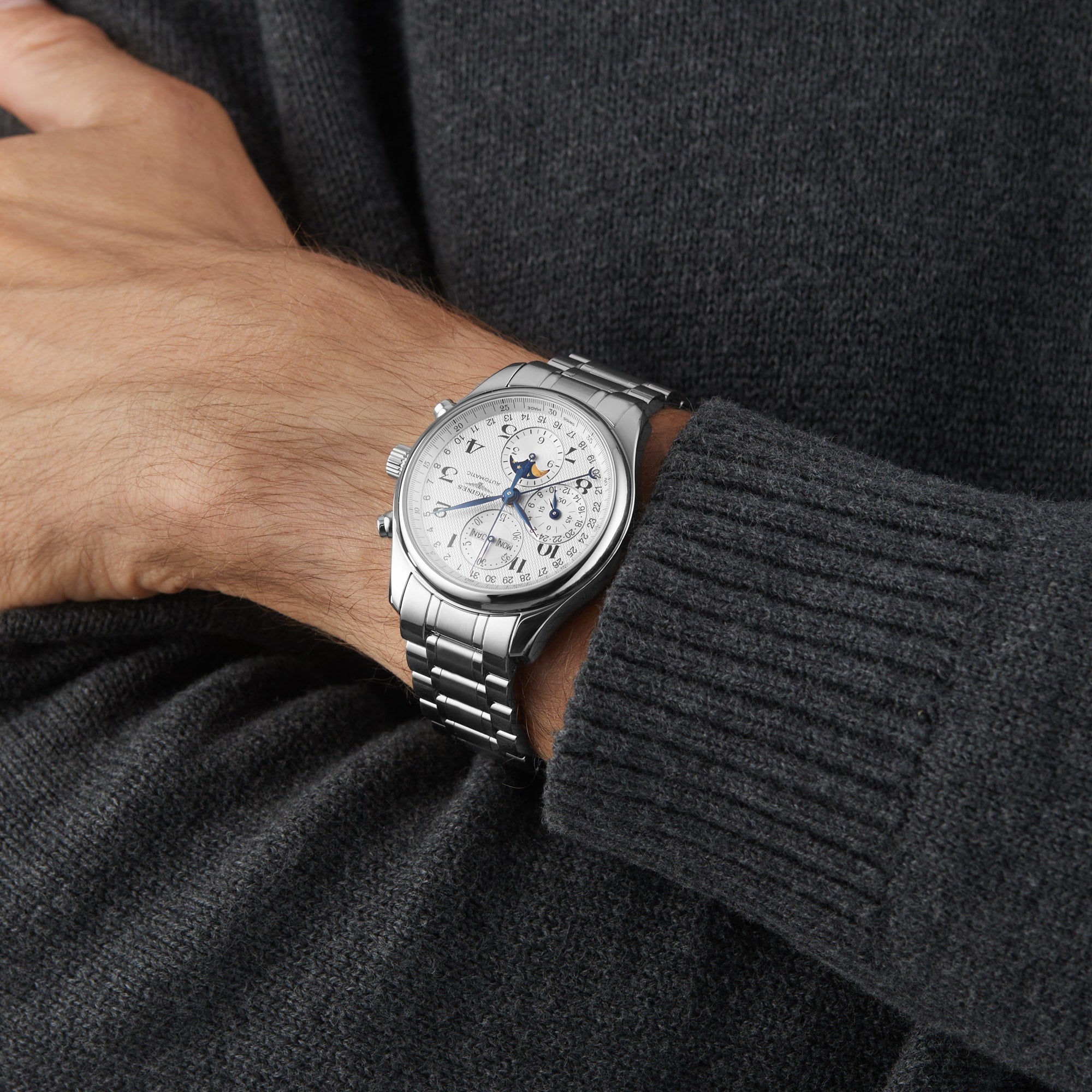 A person in a dark knit sweater wears a Longines Master Collection Chrono Moonphase silver analog wristwatch with a moonphase display and metal bracelet.