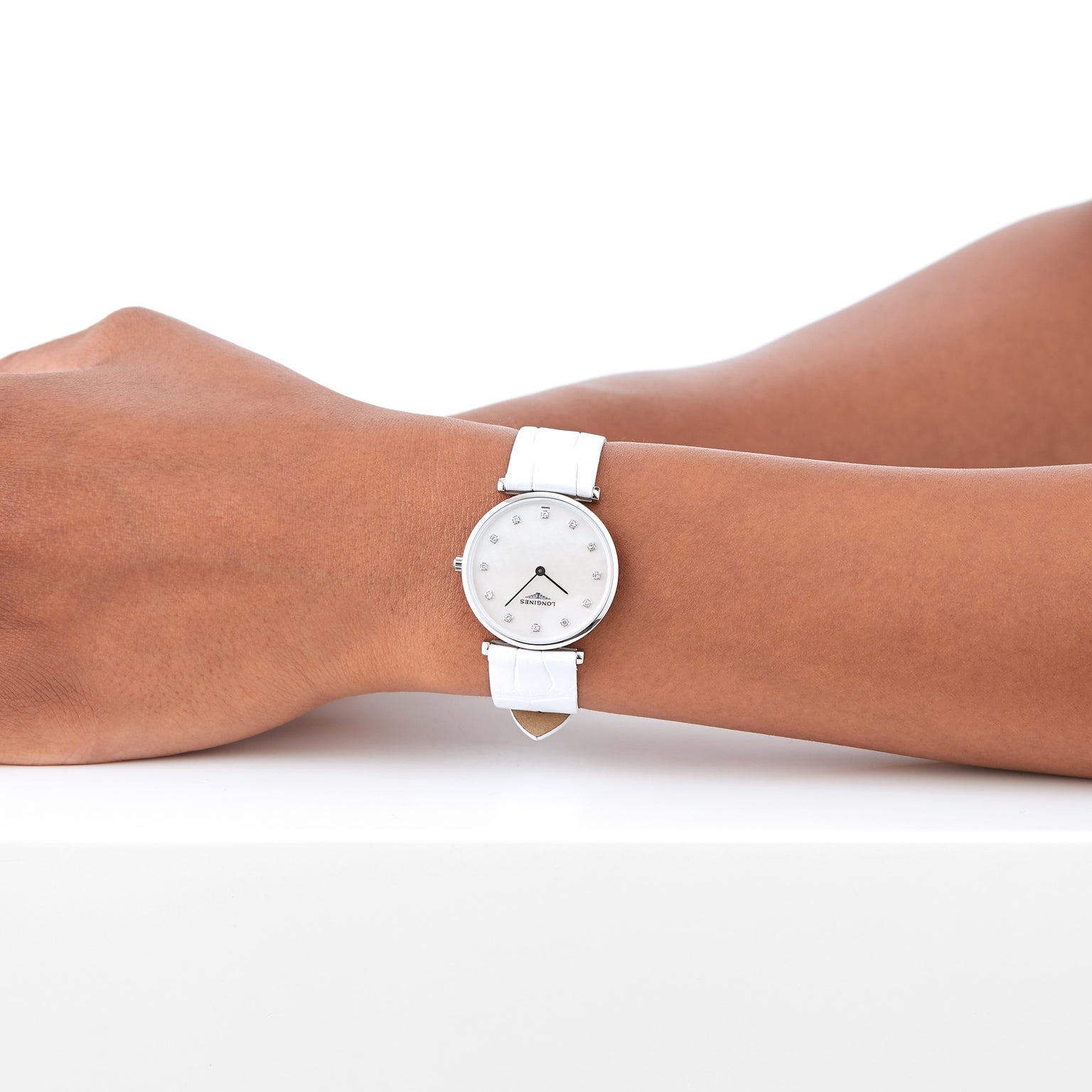 A person wears a Longines La Grande Classique de LONGINES wristwatch with a white strap, silver casing, and white mother-of-pearl dial, resting their arm on a white surface.
