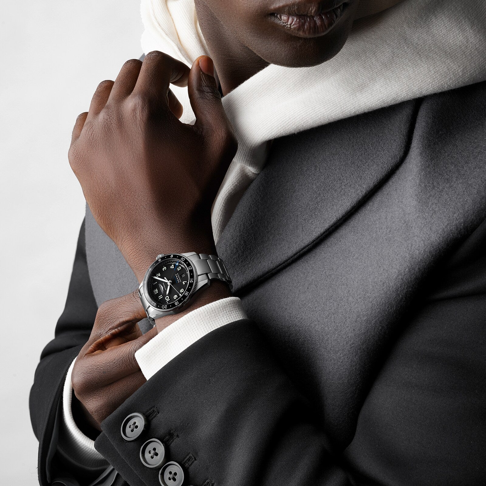 A person in a grey suit and white hoodie wears a Longines Spirit Zulu Time watch with a stainless steel case and black face on their left wrist.