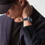 A person in a dark blazer and cap looks down, one hand touching the back of their neck, showcasing a Longines Spirit Zulu Time on their wrist.