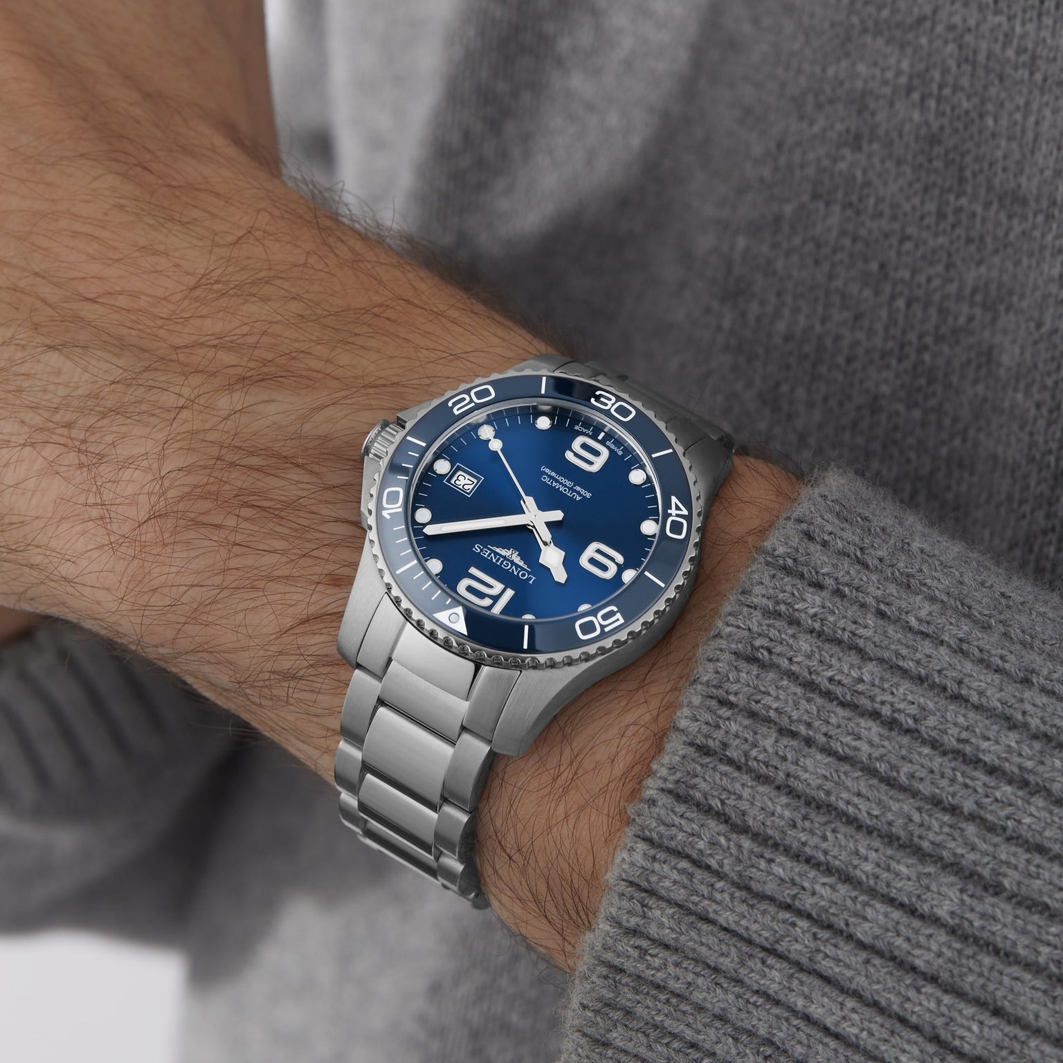 A person wearing the Longines HydroConquest, featuring a sunray blue dial with large white numerals and a stainless steel bracelet, over a gray knitted sweater.