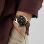 A person in a brown long-sleeve shirt and beige pants wears a Longines Spirit Zulu Time watch with a black and gold face on their left wrist, capturing the spirit of exploration.