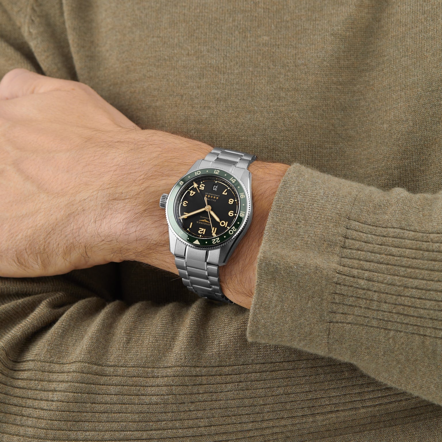 A person in a brown sweater shows off a Longines Spirit Zulu Time watch with a black dial, yellow numerals, green ceramic bezel, and stainless steel case on their left wrist.