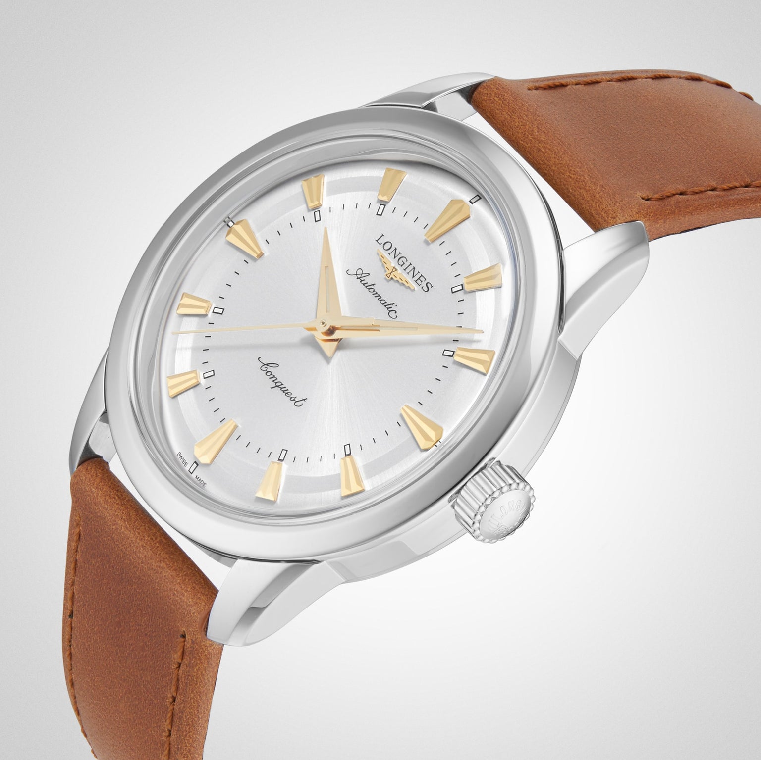 The Longines Conquest Heritage, a silver wristwatch with a white dial, gold hour markers, and brown leather strap, is shown at an angle on a light background—capturing classic Swiss watch precision.