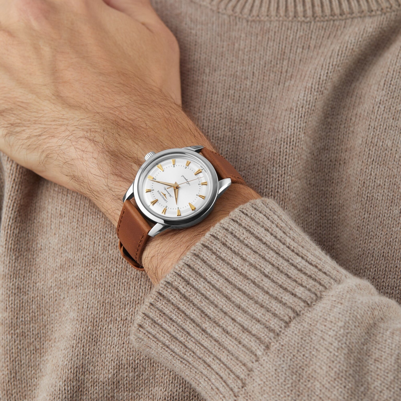 A person wears a beige sweater with a silver Longines Conquest Heritage watch featuring a white dial, gold markers, and a brown leather strap—a Swiss timepiece celebrated for its accuracy.