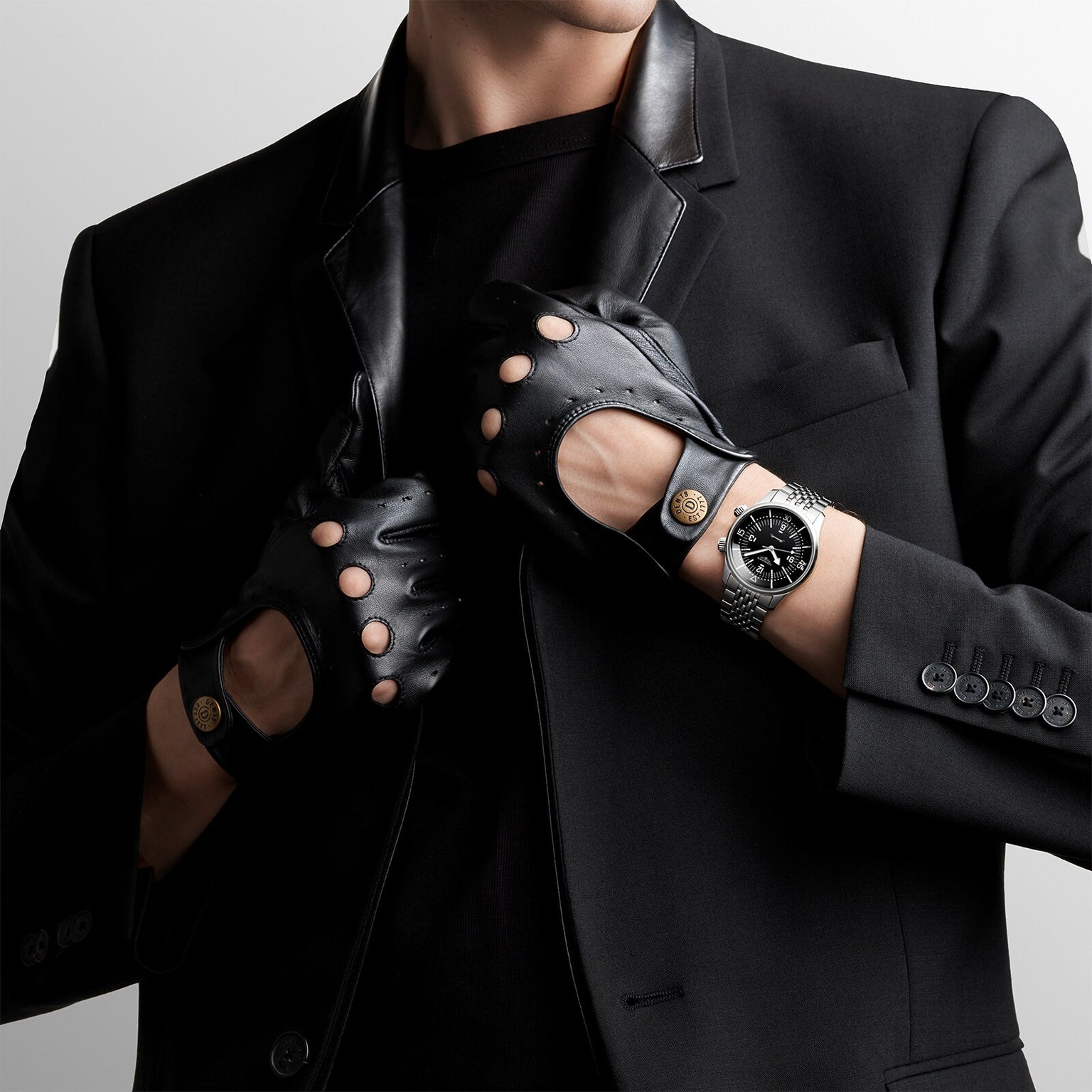 A person in a black suit and cutout leather gloves adjusts their jacket collar, showcasing the Longines Legend Diver watch on their left wrist.